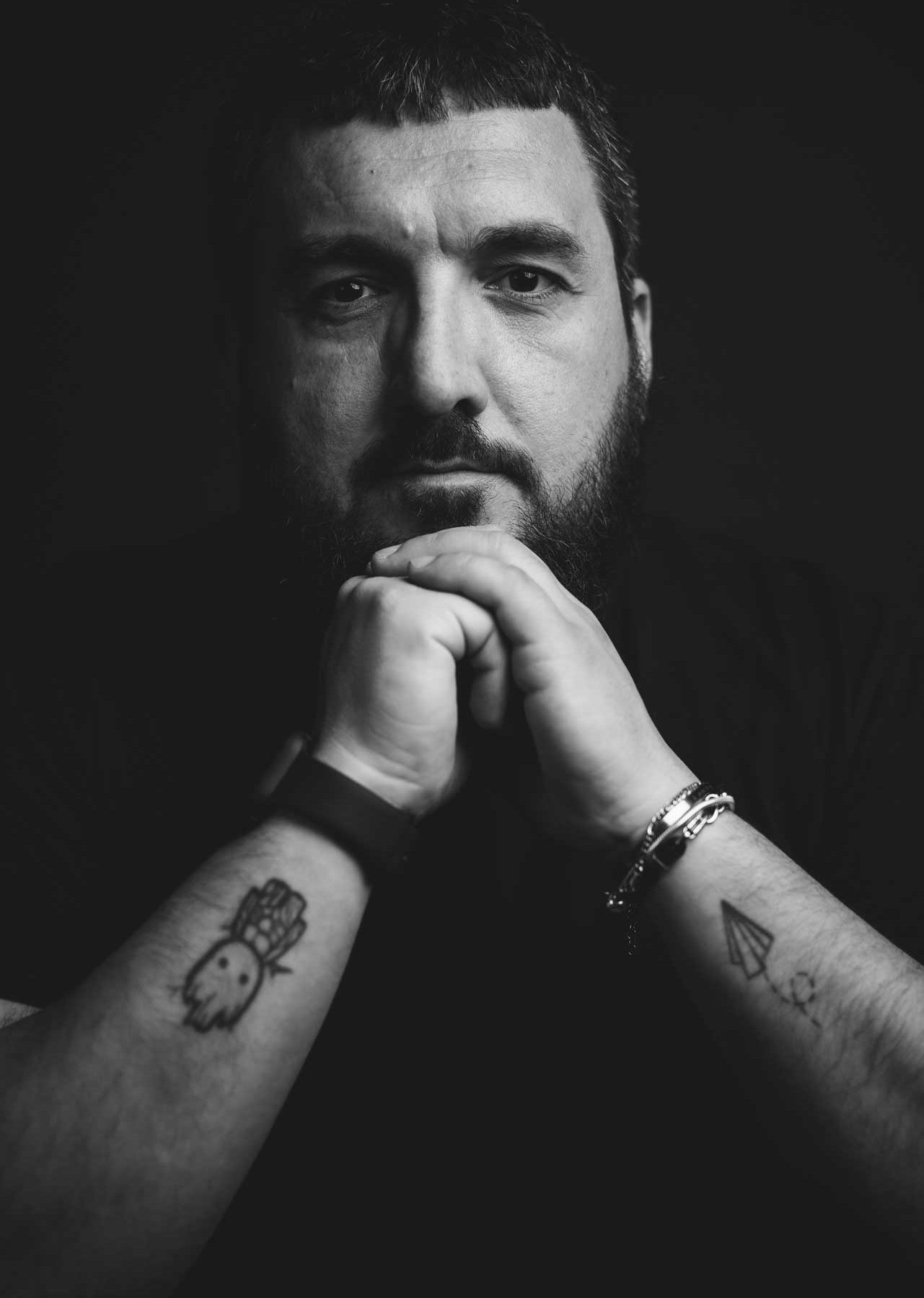 Primo piano in bianco e nero di Max Giubilei, fotografo, con le mani giunte e tatuaggi visibili